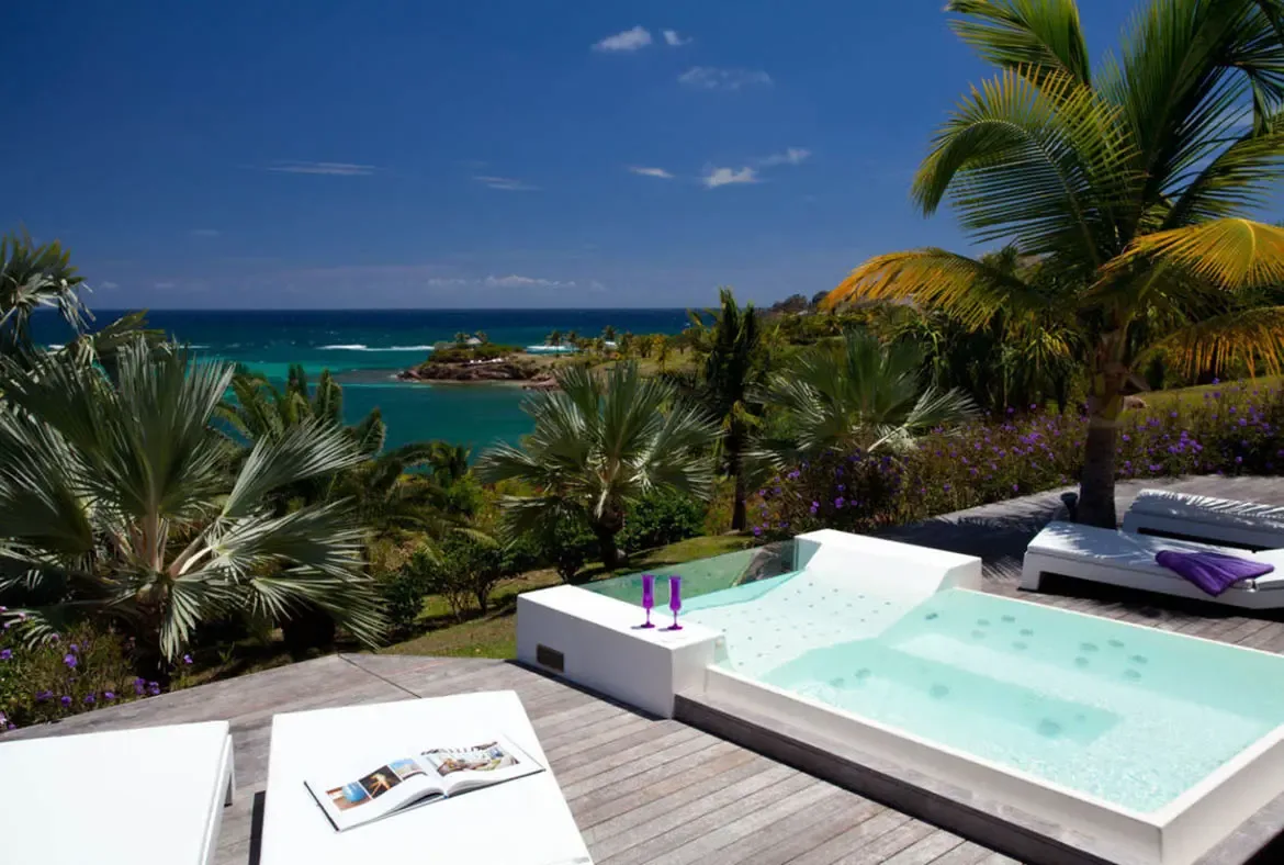 Purple champagne flutes rest on the edge of a white plunge pool, where loungers and purple flowers border a deck overlooking turquoise water and a distant green peninsula.
