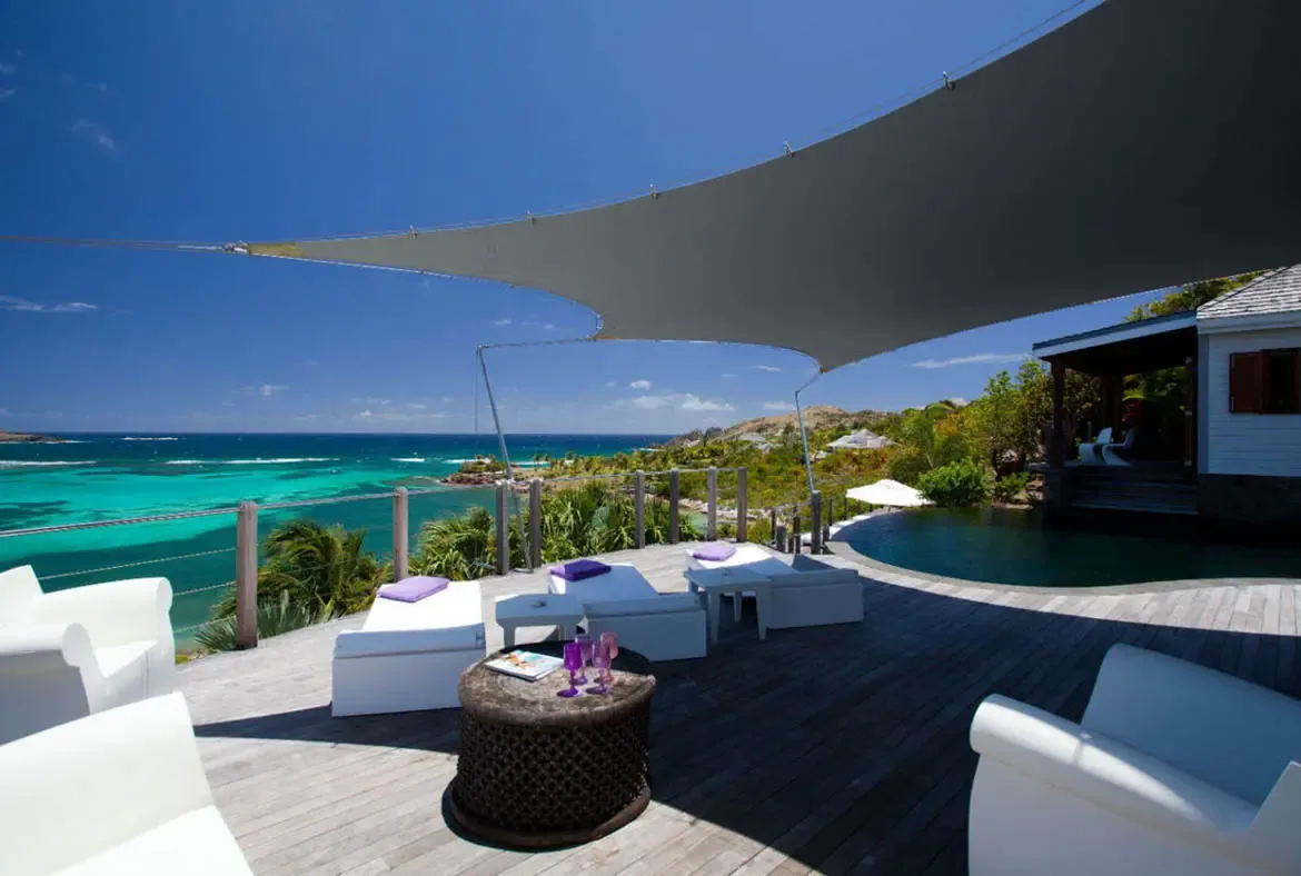 White seating with purple pillows and a sail shade sit on a weathered deck, where a dark infinity pool curves toward a turquoise bay and green hills dotted with white villas.