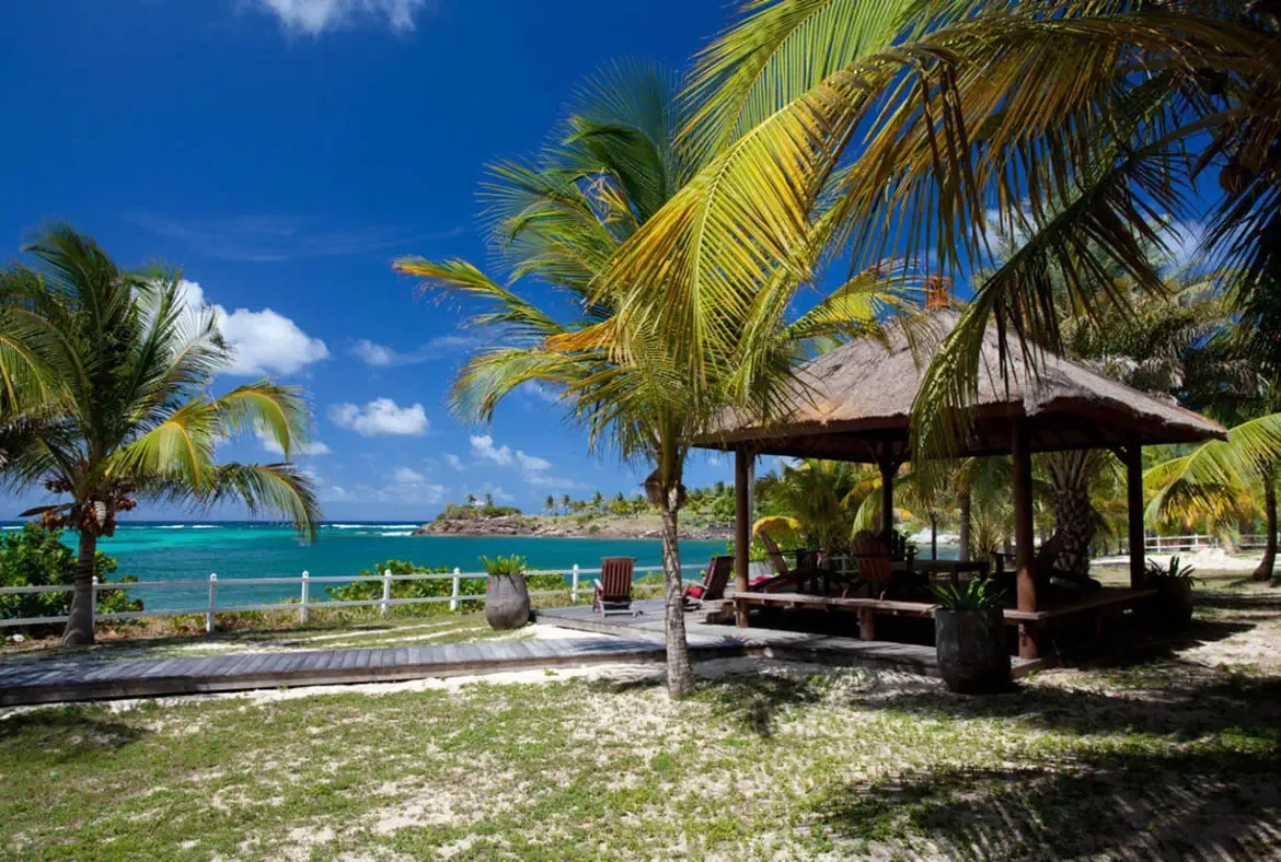 A peaked thatched gazebo sits on a wooden deck above lounge chairs, where a boardwalk through palm trees leads to turquoise water and a green peninsula on the horizon.