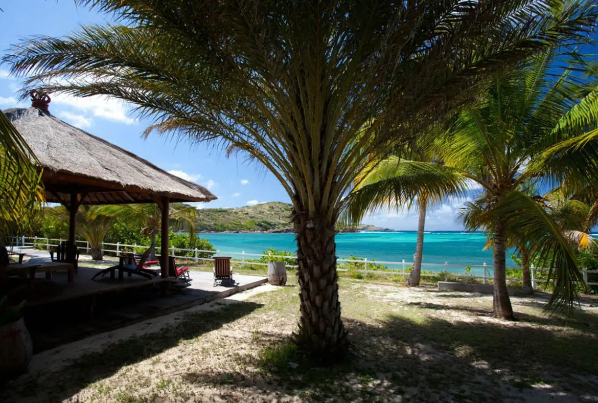 A traditional thatched pavilion and palm fronds frame a sandy garden path, where a white fence and turquoise bay create a secluded beachfront paradise.