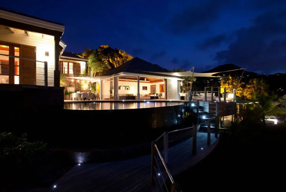 At blue hour, a dark infinity pool reflects warm villa light while tiered wooden decks and white sail canopies connect indoor living to a rocky hillside lit with golden uplights.