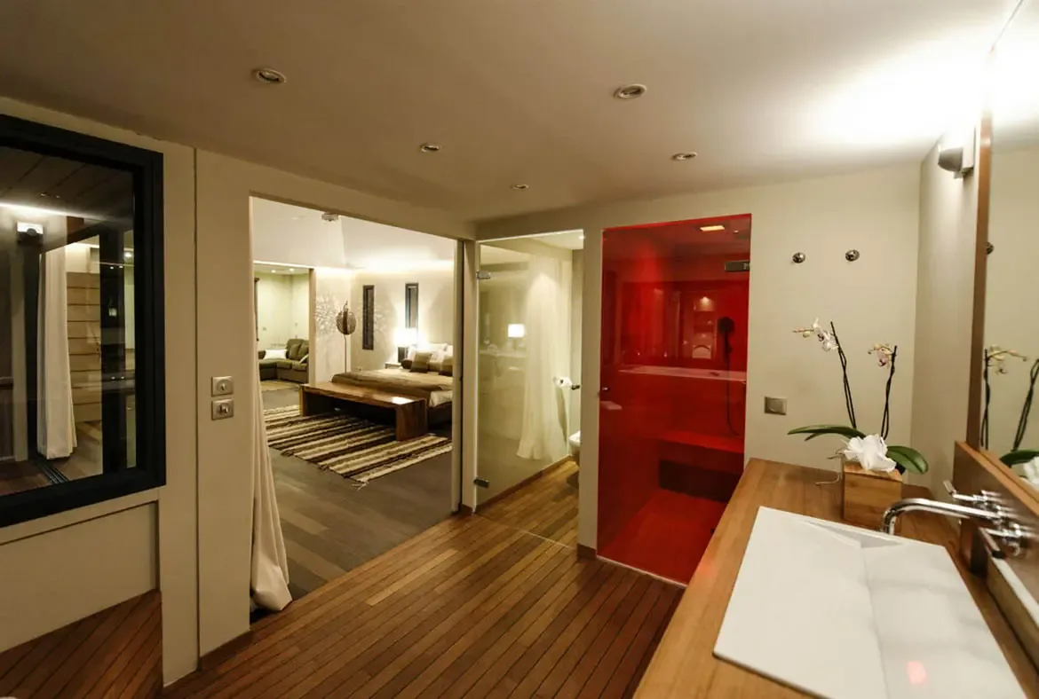 A striking translucent red glass shower steals the spotlight in this open-plan suite, while a warm wood vanity with orchid and a view into the bedroom beyond keep the mood polished and serene.
