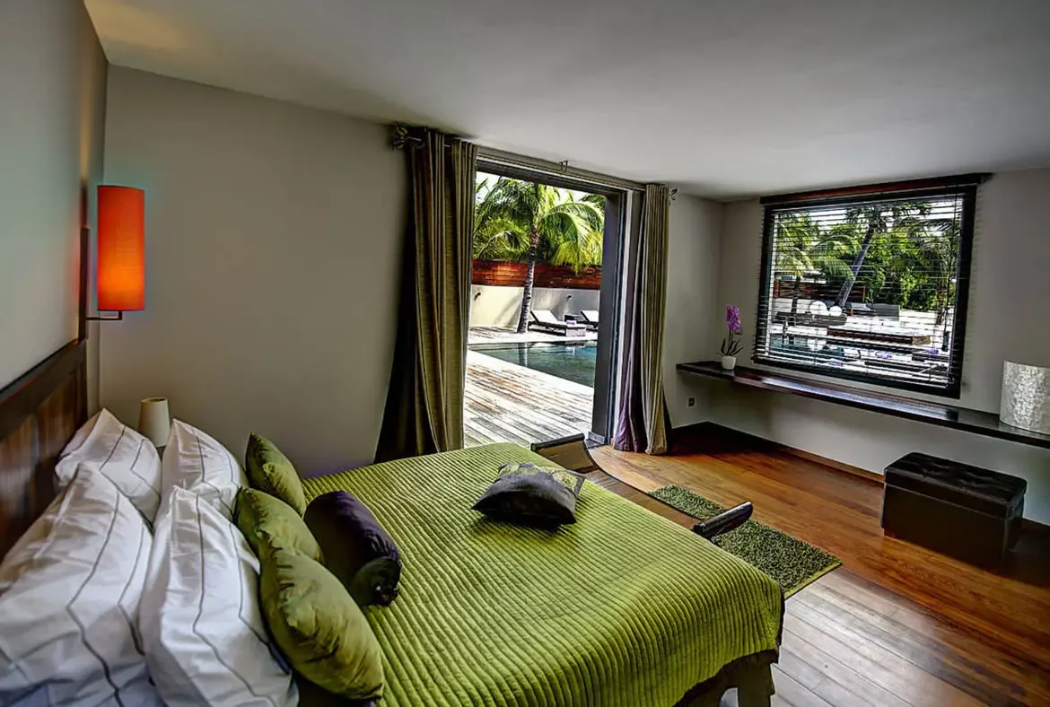 A lime-green bedspread and warm orange sconce bring personality to this bedroom, while open doors lead straight to the turquoise pool and palm-lined deck beyond.