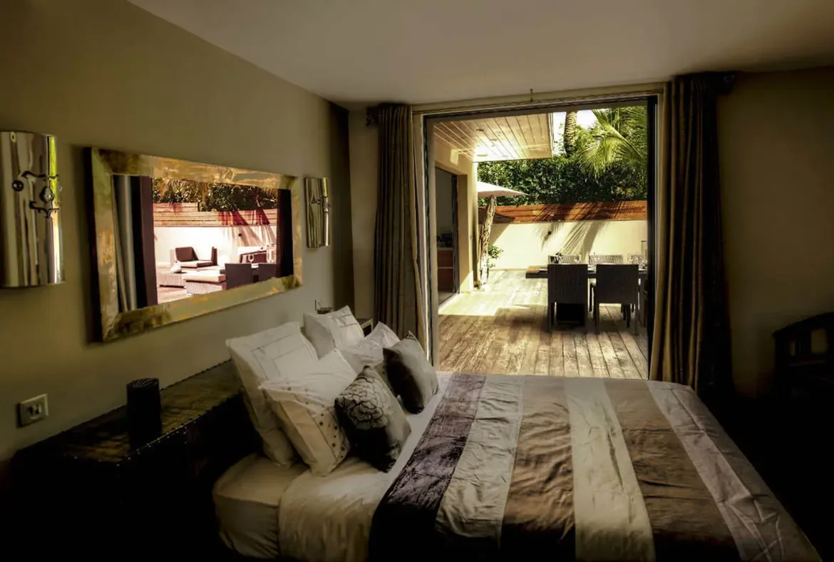 Wide-open glass doors connect this earthy bedroom directly to a shaded terrace dining area, with a large mirror reflecting the greenery outside. The indoor-outdoor flow feels effortless and tropical.