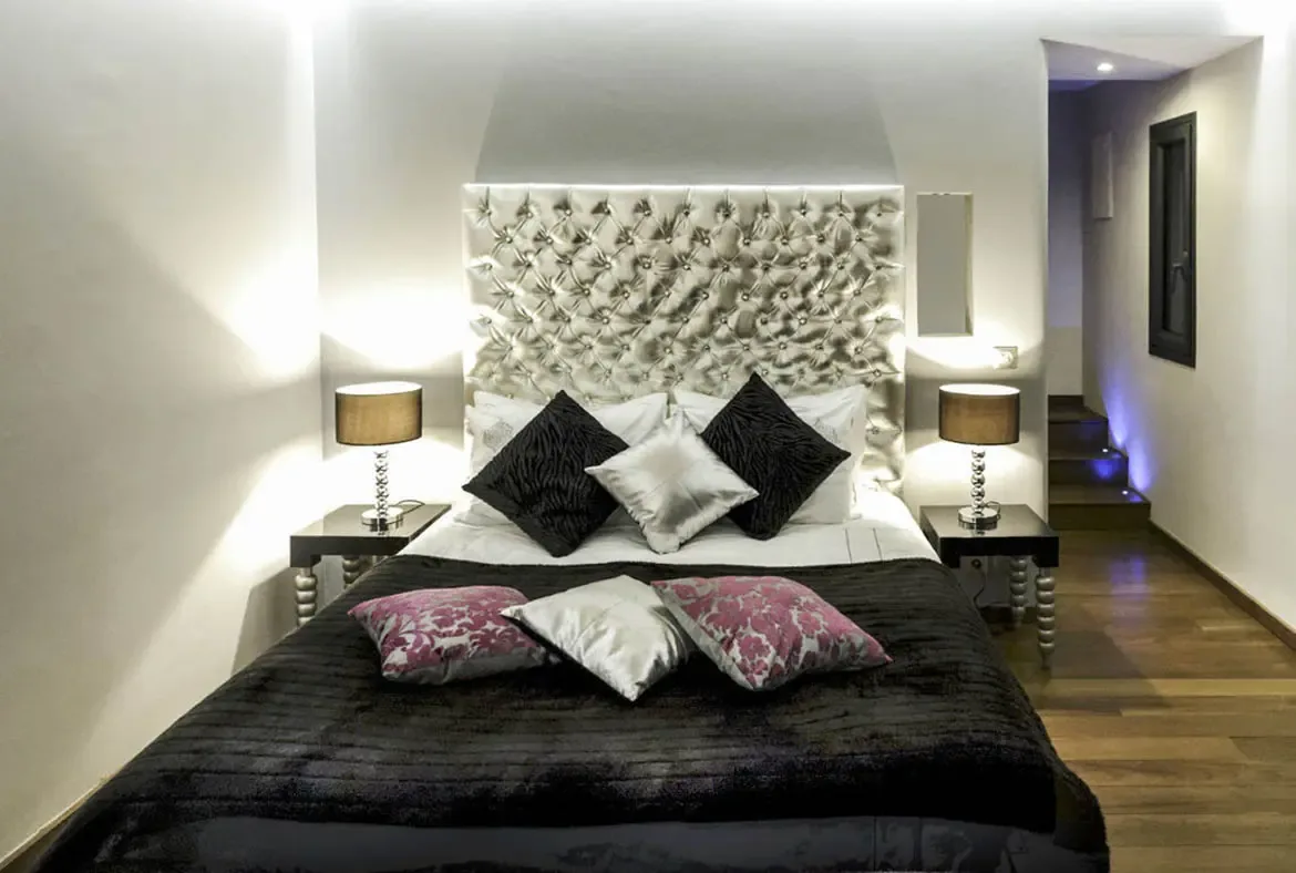 A shimmering silver tufted headboard and magenta floral pillows give this bedroom a glamorous edge. Blue-lit stairs in the background add a cool, contemporary contrast to the warm bedding.