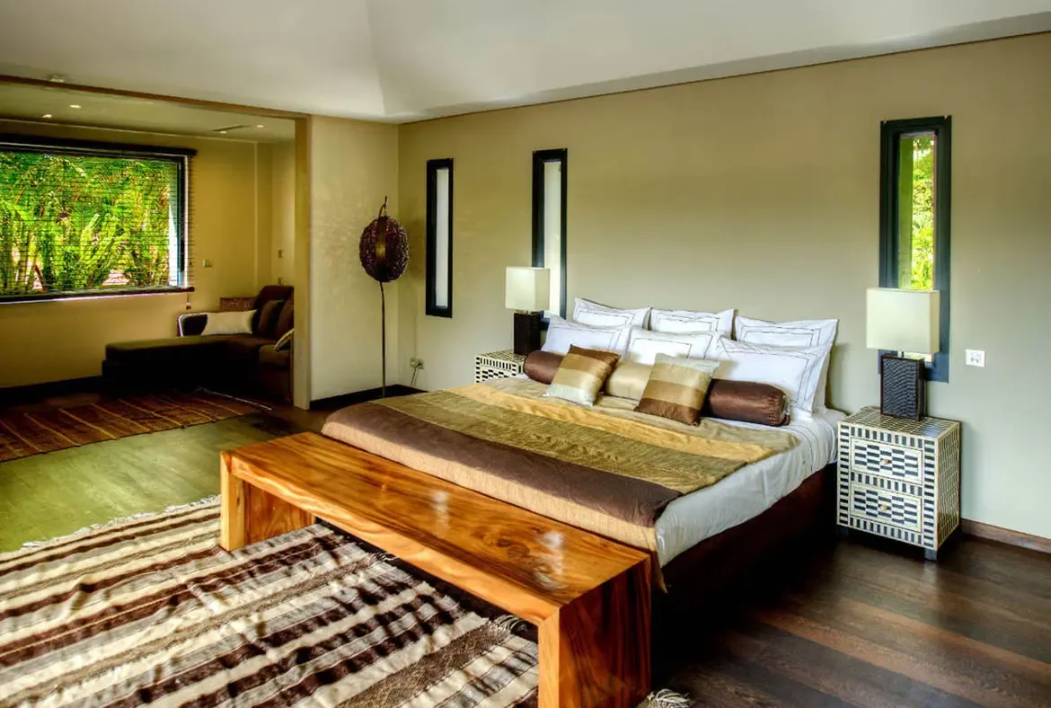 Crisp white bedding, a polished live-edge bench, and geometric bone-inlay nightstands bring warmth to this vaulted-ceiling bedroom. A lounge nook overlooks lush greenery through a wide window.