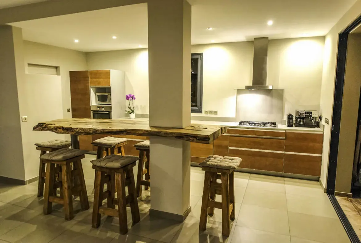 A thick live-edge wood bar counter with chunky stools is the centerpiece, backed by sleek cabinetry, a gas cooktop with professional hood, and an espresso machine. Rustic meets contemporary in the best way.