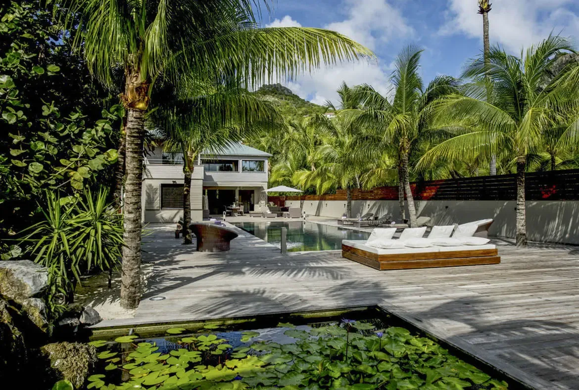 A lily pond with purple blooms sits at the edge of the deck, while a white-cushioned daybed, turquoise pool, and the villa's grey facade lead the eye to the green mountain beyond.