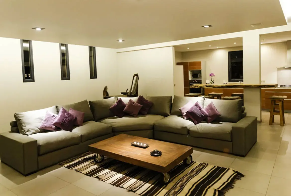 A muted olive-gray sectional with lavender pillows anchors this warm living room, paired with a live-edge wood coffee table on casters. The space flows into a kitchen with a natural-wood breakfast bar.