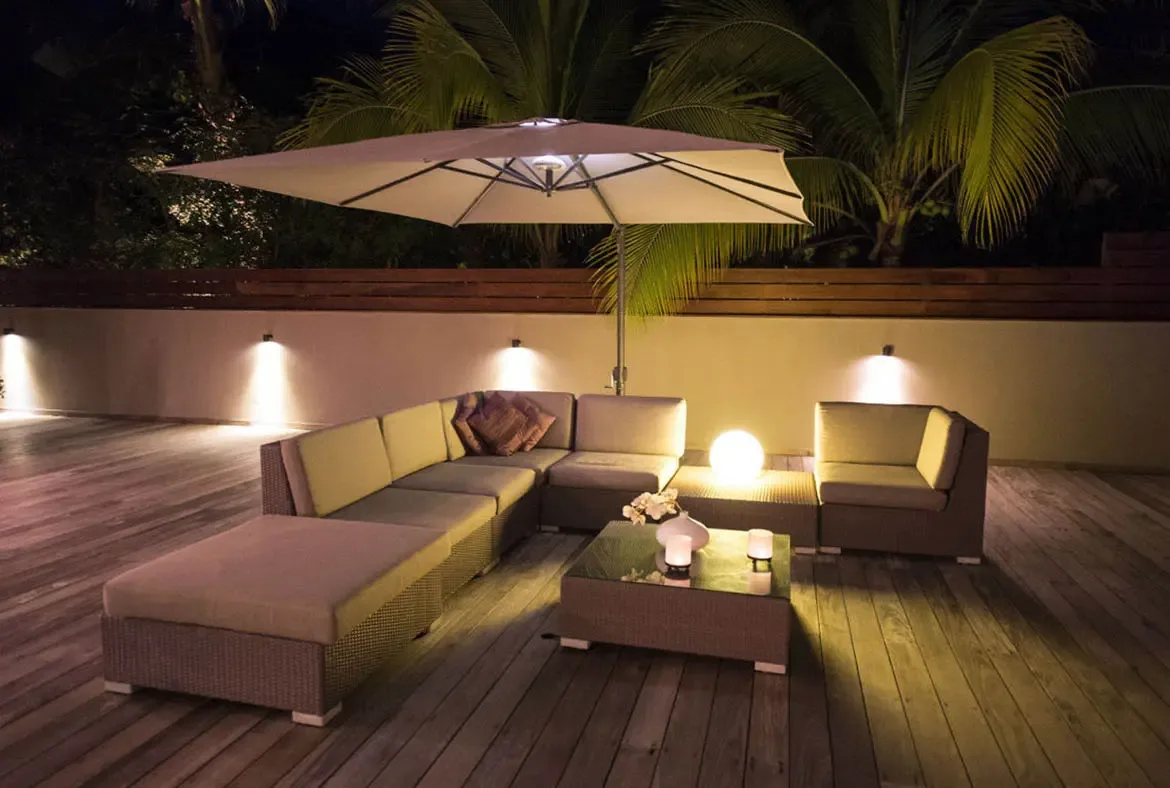Under a glowing rectangular patio umbrella, a cream-cushioned sectional and a warm sphere lamp create the perfect evening lounge on the wooden deck, framed by uplit palms and a tropical sky.