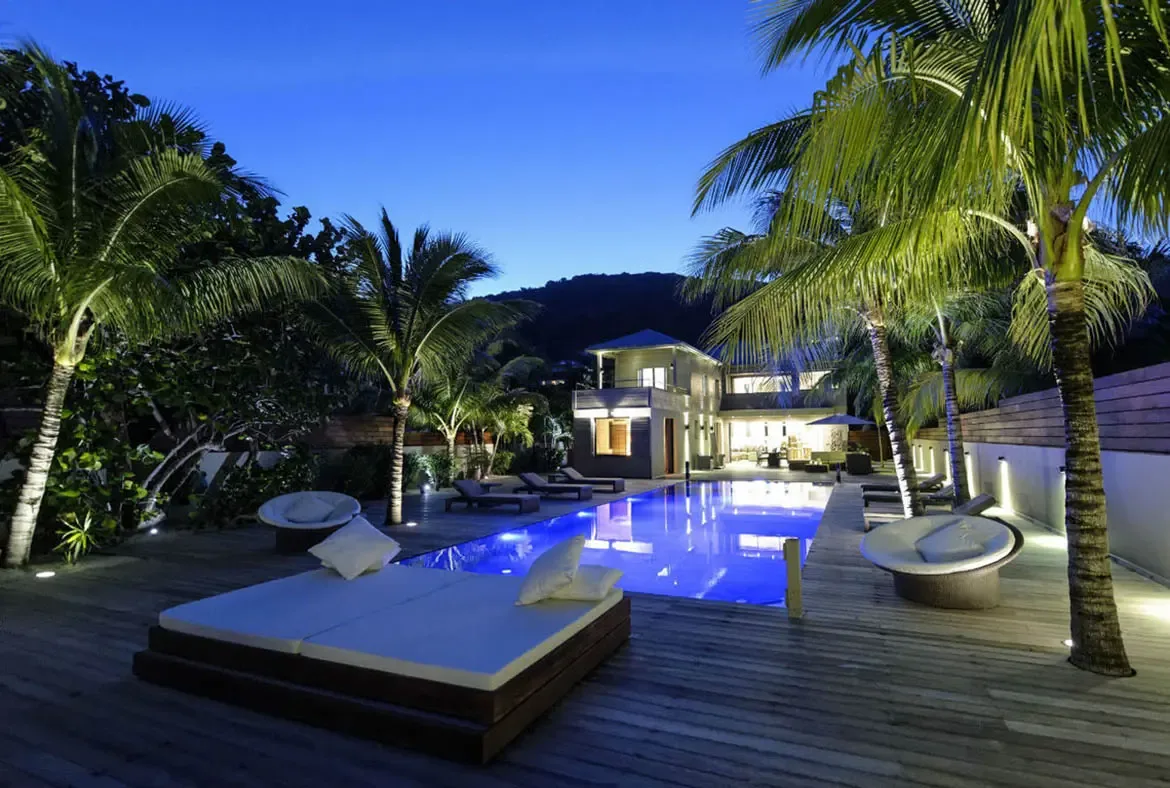 A plush white daybed sits poolside under dramatic uplit palms, while the two-story villa glows behind. The cool blue water and warm lighting create a cinematic tropical night.
