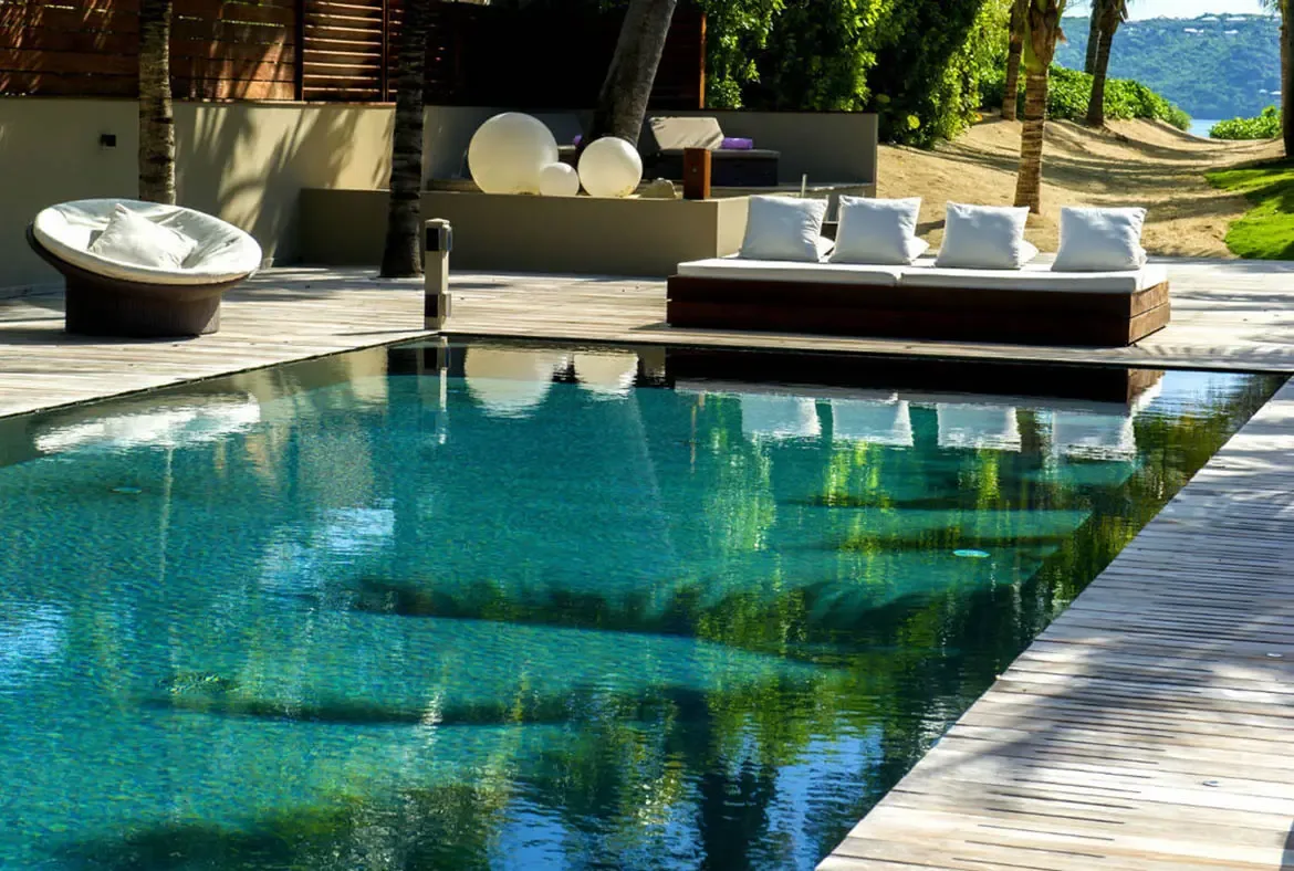 A long turquoise pool is flanked by a circular wicker daybed and a rectangular cushioned lounger at the water's edge, with a sandy path leading toward a distant blue bay through the palms.