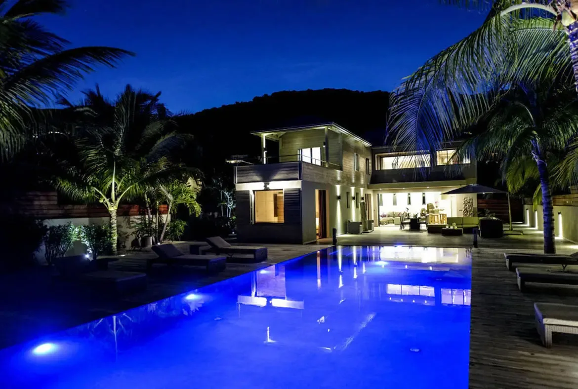 At night, the villa's open-concept living and dining area spills onto the deck beside a glowing rectangular pool. Warm lights and tall palms make the whole space feel like a private evening retreat.