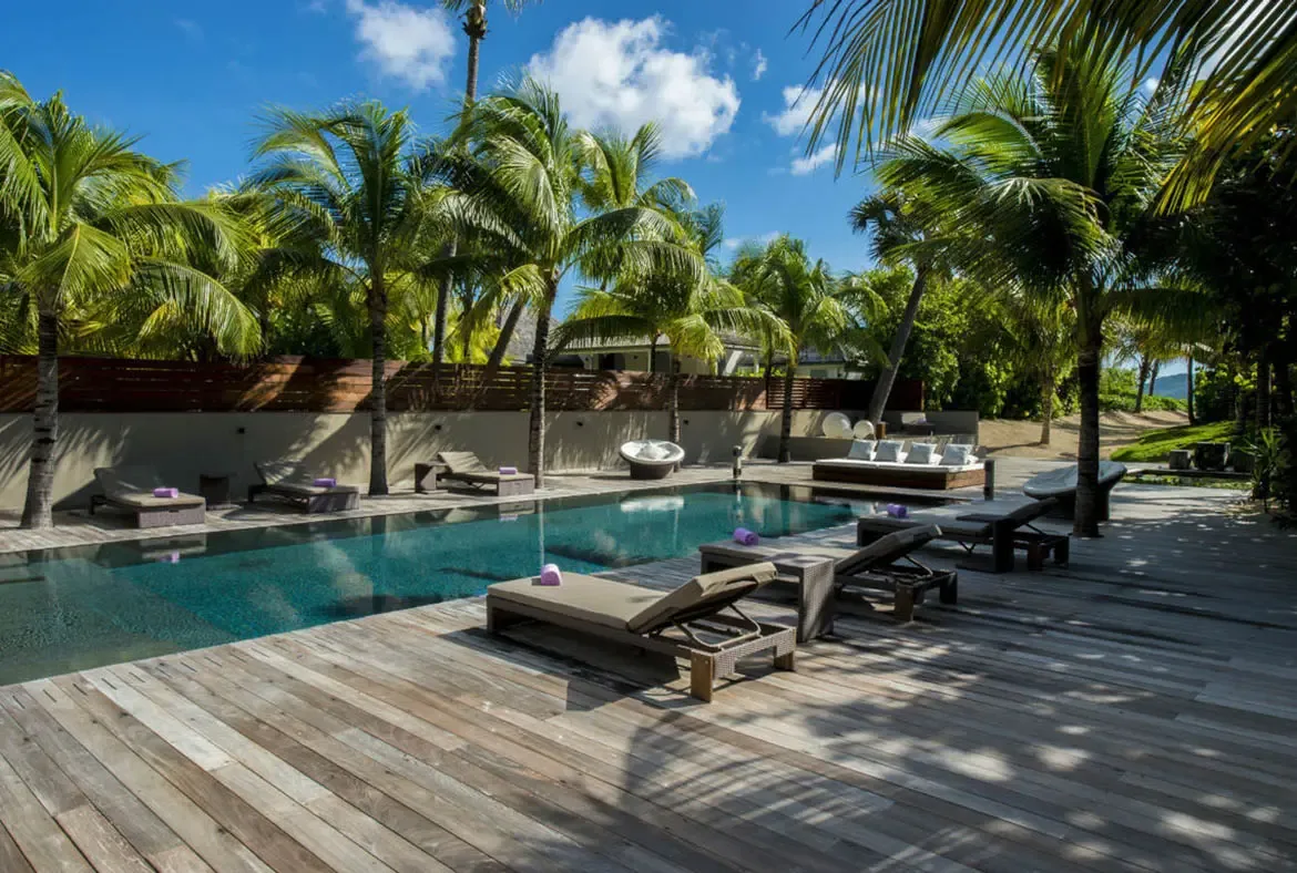Dappled palm shadows play across the weathered deck surrounding the turquoise pool, where loungers, a white bowl chair, and a plush daybed invite full-day relaxation under the canopy.