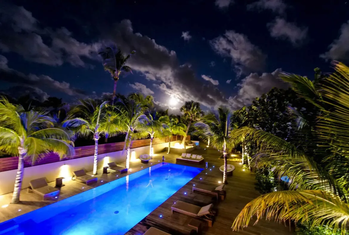 Under a moonlit sky with swirling clouds, the villa pool glows deep blue while warm sconce lights and uplit palms add drama to this magical Caribbean night.