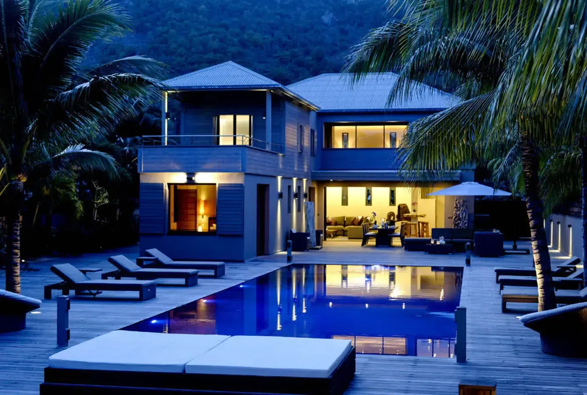 Modern two-story villa glows at dusk, with a luminous pool reflecting warm interior light through wide-open doors. Palm trees and a steep hillside frame this serene twilight scene.