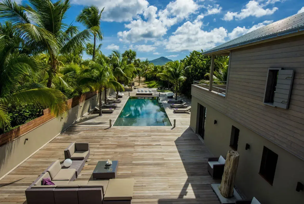 From above, the turquoise pool stretches toward a green mountain, flanked by an L-shaped outdoor sofa, purple-cushioned loungers, and the villa's contemporary wood-sided facade.