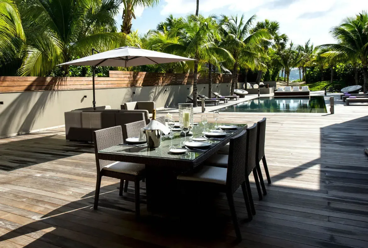 An elegant table set for six with champagne on ice sits on the poolside deck, while lounge chairs with purple cushions and a white umbrella lounge complete the alfresco scene.