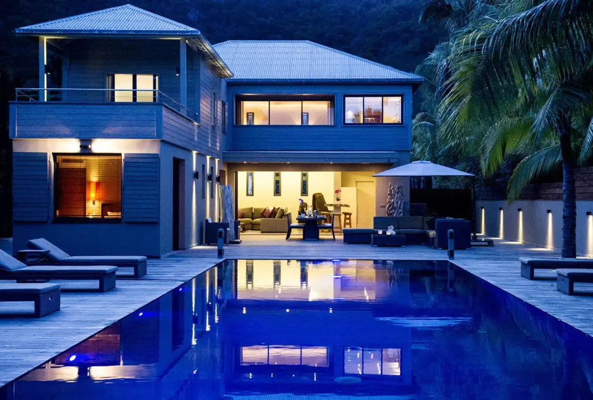 The modern grey-sided villa opens wide at dusk, with an outdoor lounge under a white umbrella and a long pool reflecting the warm interior. A green hillside rises behind for total privacy.