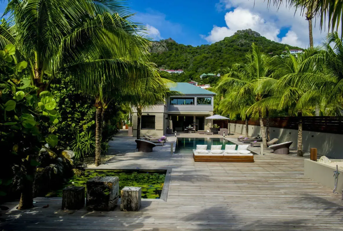 A contemporary two-story villa rises against a lush green mountain, with a long pool, a lily-pond accent, and varied lounge areas spreading across the weathered wooden deck.