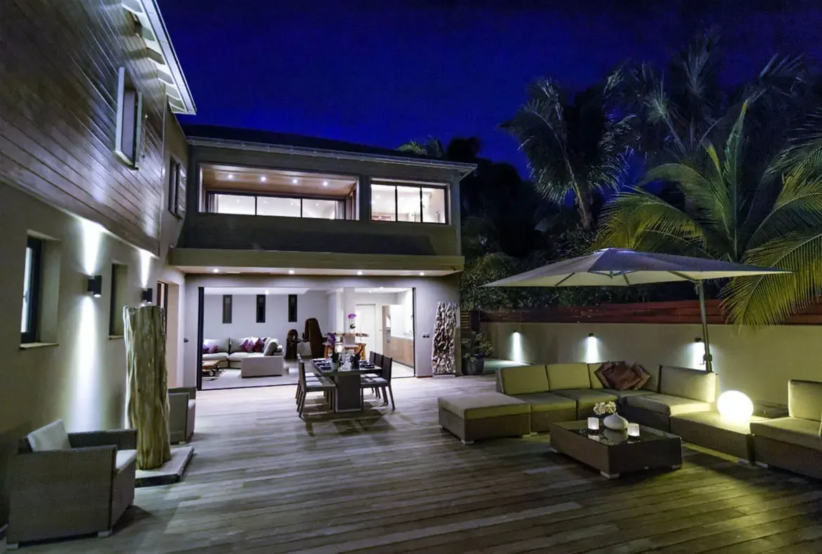 At twilight, warm uplighting and a glowing globe lamp transform the covered dining and lounge terrace into an intimate evening space, with the villa's bright interior visible through open glass walls.