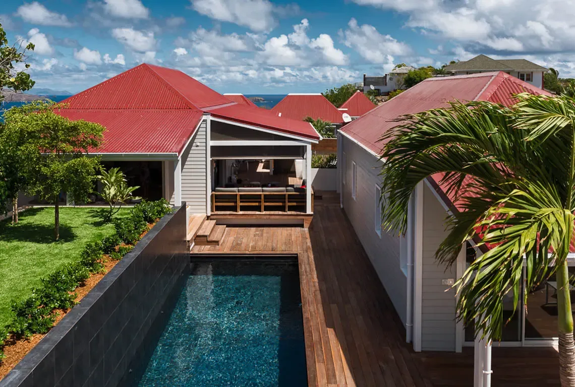 An aerial view reveals the villa's pavilion layout connected by wooden decking. The dark-tiled lap pool anchors the courtyard, while red corrugated roofs crown each structure. Manicured lawns, tropical landscaping, and ocean glimpses create a private hillside oasis.