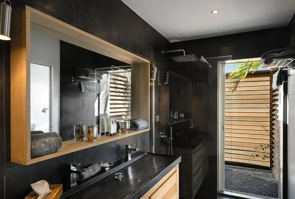 Dark grey tile walls and a black vanity with chrome fixtures create a sleek modern bathroom. A wood-framed horizontal mirror, walk-in glass shower, and glimpse of tropical greenery through the door blend contemporary design with island freshness.