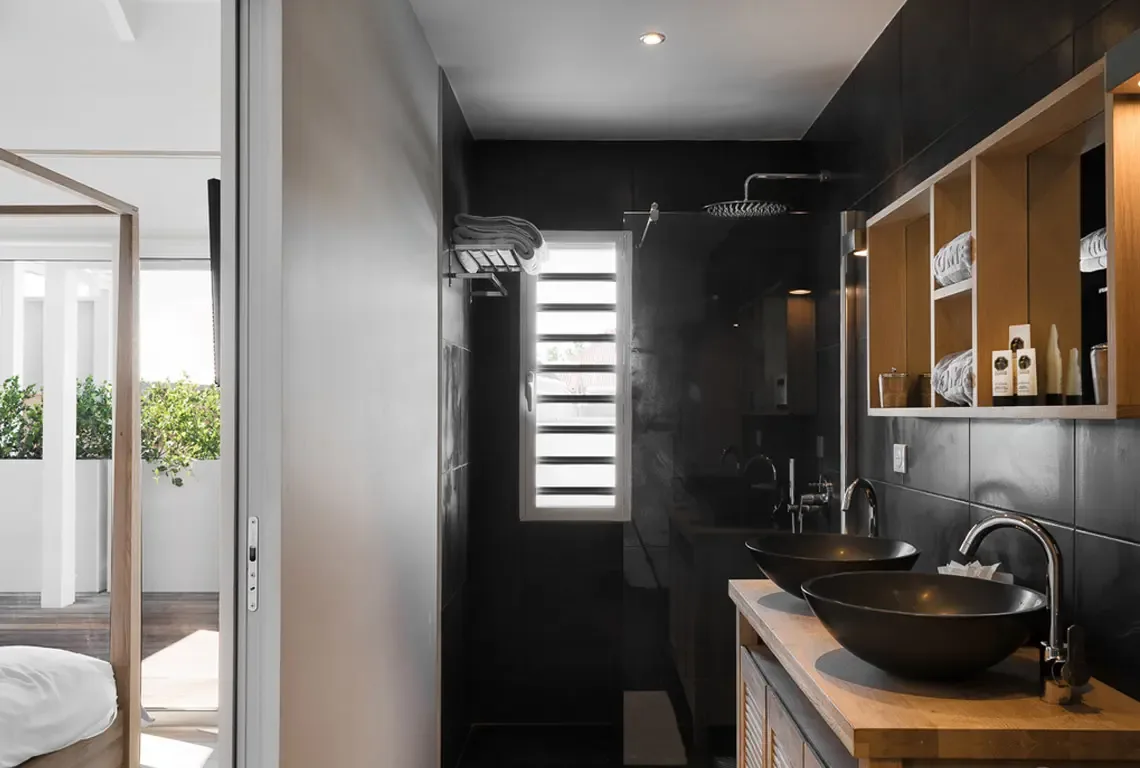 Dark glossy tile walls frame this spa-like bathroom where a wooden vanity holds two black vessel sinks with chrome faucets. Open shelving displays neatly arranged toiletries, while a horizontal slatted window and glass-enclosed rainfall shower complete the sophisticated retreat.