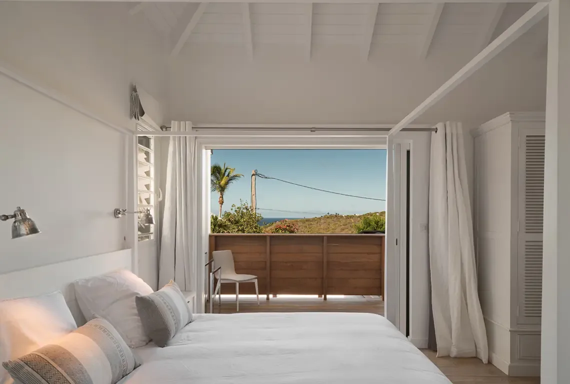 White curtains frame sliding doors that open to a private balcony with ocean horizon views. A king bed with striped pillows rests beneath vaulted white-beamed ceilings, while an industrial wall lamp and white louvered wardrobe complete this bright, airy retreat.