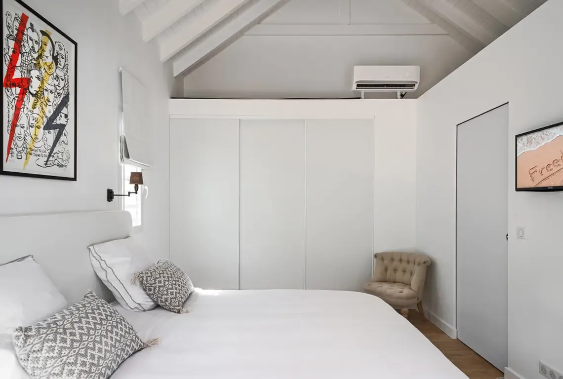 White vaulted-beam ceilings soar above this minimalist bedroom where a king bed with geometric grey-and-white pillows anchors the space. A bold colorful artwork with lightning bolts adds vibrant personality, while a beige tufted armchair and white sliding closets complete the serene retreat.