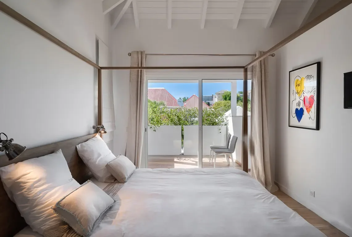 Crisp white linens and layered pillows dress a king bed with sleek dark wood headboard. Sliding glass doors open to a balcony with two white chairs and red-roofed views, while a playful framed artwork adds personality—serene indoor comfort meets outdoor Caribbean living.