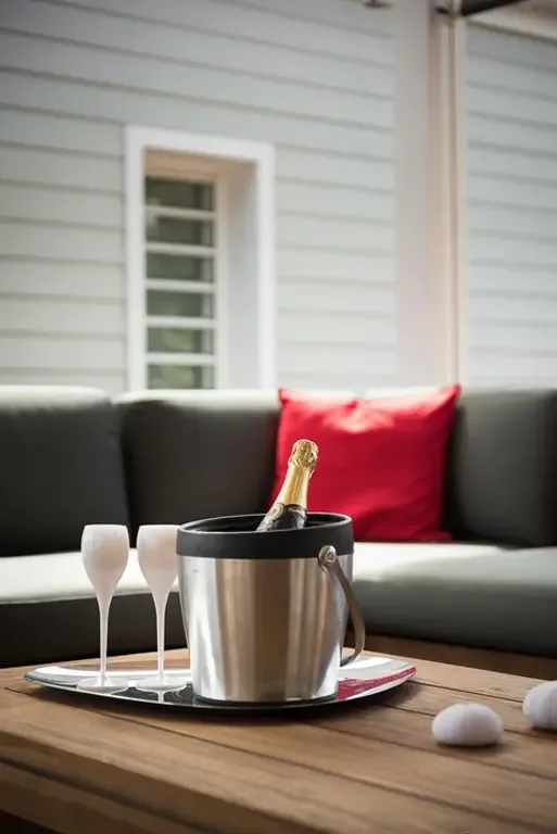 A chilled bottle of champagne rests in a stainless steel ice bucket beside two tall white glasses on a wooden table. Behind, a grey sofa with red pillow sits before white horizontal slats—relaxed elegance awaits your welcome toast.