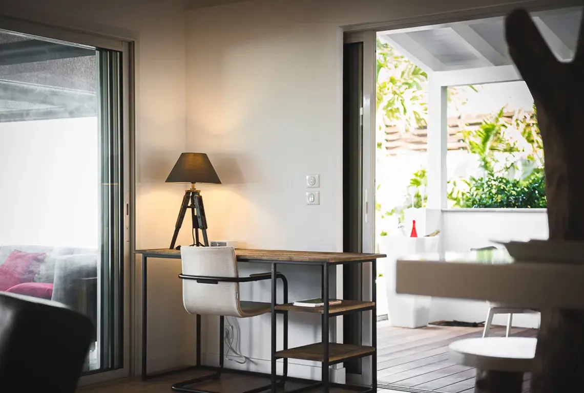 A compact desk with black-shaded lamp sits between two sliding glass doors, one opening to a balcony with green planters. A white chair and wooden shelf complete this functional workspace where tropical views inspire productivity.