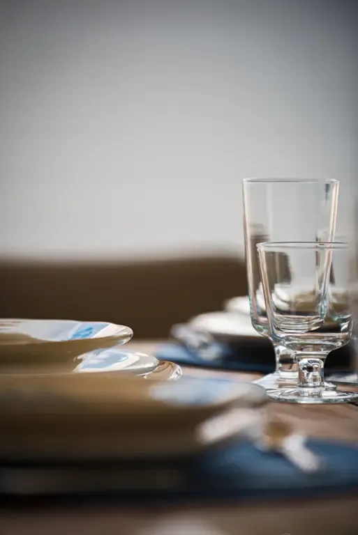 Stacked plates with subtle blue patterns and crystal glassware rest on a blue placemat. Soft natural lighting and shallow depth of field create an intimate, refined atmosphere—every detail curated for an elegant dining experience.