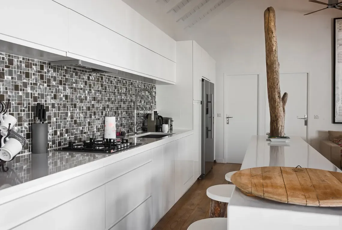 A sleek white kitchen with metallic mosaic backsplash and gas stovetop stretches along one wall. The center island features a striking natural tree sculpture in a white pot, while three barstools and light wood floors complete this modern culinary space beneath a slanted white ceiling.