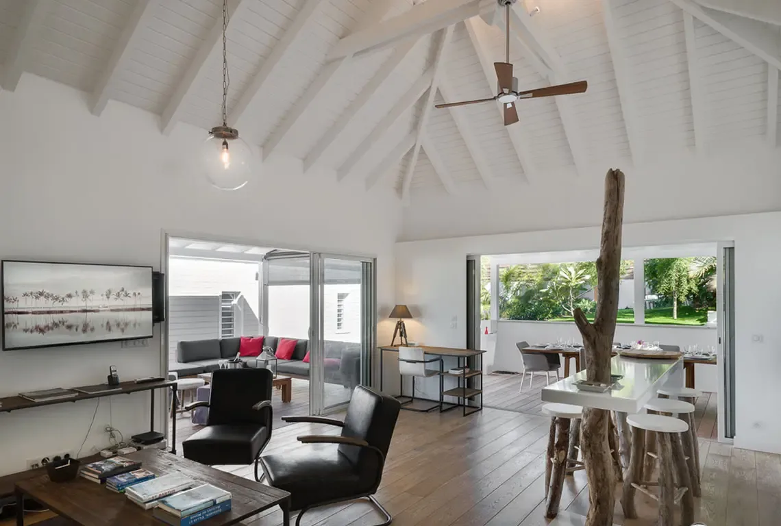 Vaulted white-beamed ceilings soar above this open-concept living space. A breakfast bar with natural tree-trunk supports anchors the room, while two black armchairs and a wall-mounted TV create a cozy lounge. Sliding glass doors open to a covered patio and dining terrace beyond.