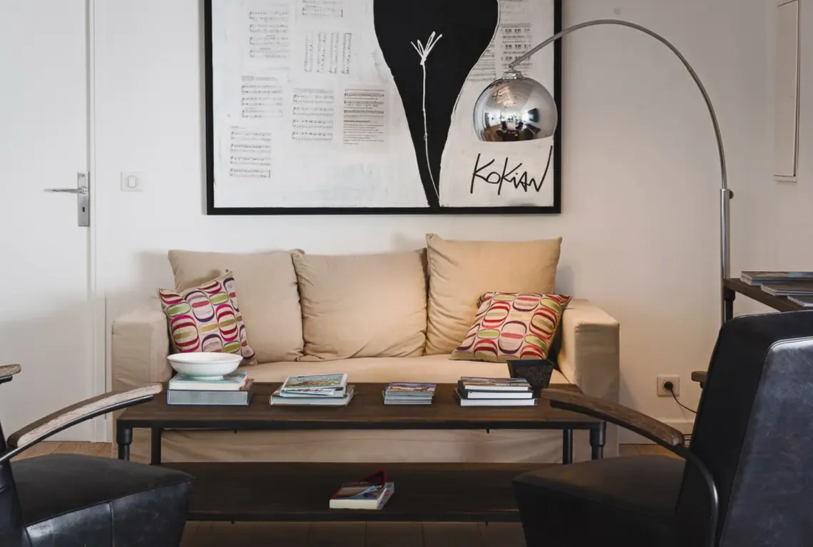 A light brown sofa with colorful cushions and two black leather armchairs face a dark wood coffee table beneath a large black-and-white abstract painting. A silver arc floor lamp and warm wood floors complete this comfortable, modern living space.