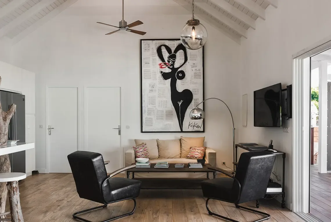 High white vaulted ceilings crown this modern living room where a large silhouette artwork hangs above a tan sofa with colorful pillows. Two black leather armchairs flank a dark wood coffee table, while a silver arc lamp and glass pendant complete the sophisticated space.