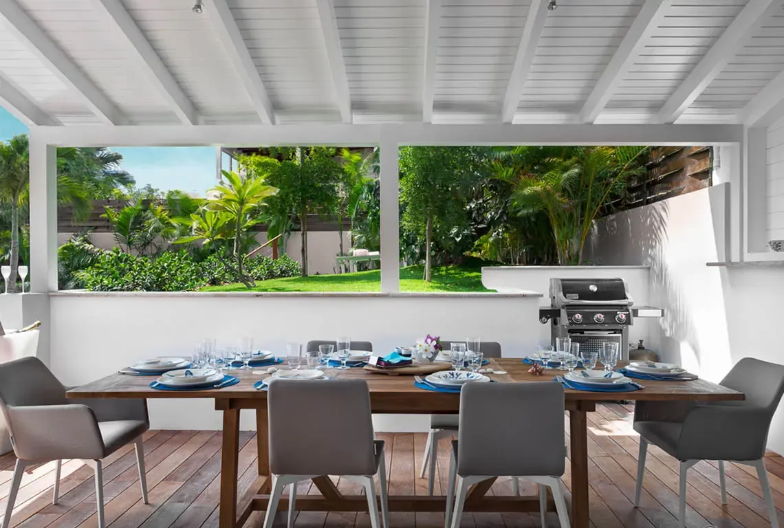 A long rustic wooden table set for six with blue and white dishware sits on a covered patio with white slatted ceiling. A stainless steel grill stands ready beside a white wall, while lush tropical gardens and palm trees frame the scene—alfresco dining in paradise.