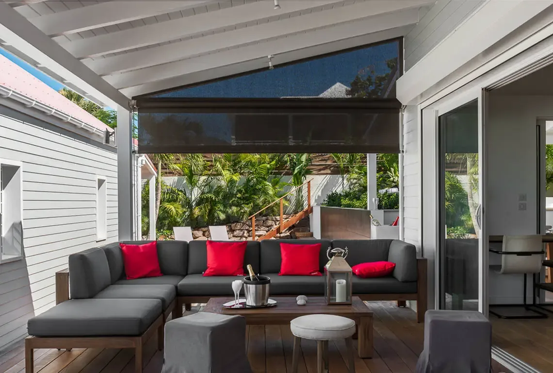 An L-shaped dark gray sofa with red pillows sits beneath a white pergola with roll-down shade screen. A low wooden coffee table holds a silver ice bucket and glasses, while gray stools and a white stool complete the seating—inviting outdoor living at its finest.