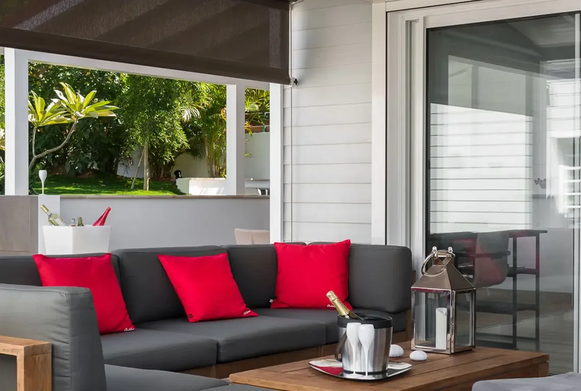 A dark grey sectional sofa with three red pillows faces a wooden coffee table holding a champagne bucket, flutes, candle lantern, and seashells. A rolled shade provides comfort while glass doors reflect the lounge—perfect for peaceful afternoons or social gatherings.