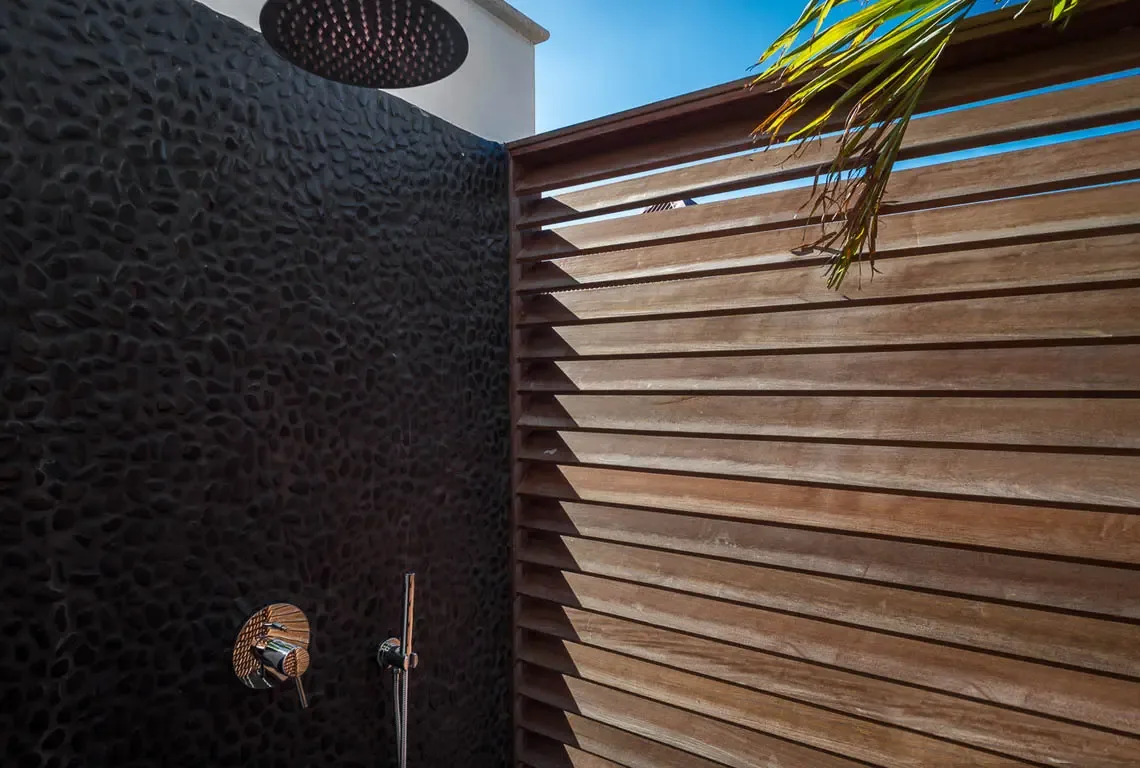 A sleek outdoor shower with black pebble-textured wall and horizontal wooden slat screen. A large rain showerhead and chrome controls promise an invigorating rinse under the open sky, while a palm frond adds tropical charm.