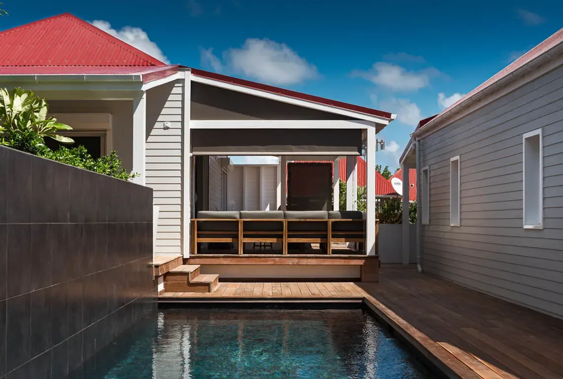 An outdoor sofa with grey cushions sits on the wooden deck beside the dark blue pool. White horizontal siding, red roofs, and a dark privacy wall frame the scene while wooden steps lead to the house—seamless indoor-outdoor living.