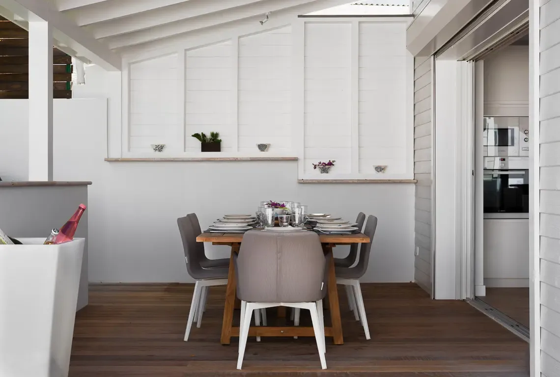 A light wood dining table with four place settings and white plates sits on a covered patio. White panel art and small plants adorn the grey wall, while an open sliding door connects to the kitchen—alfresco dining just steps from the chef.