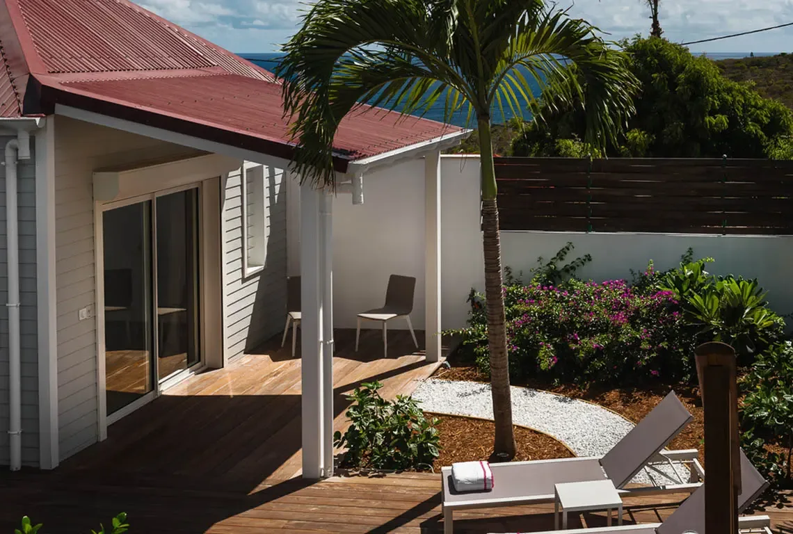 Two white loungers with pink-patterned cushions sit on a wooden patio beside a pink-roofed pavilion. Pink and green tropical flowers frame the scene, while the ocean and blue sky stretch beyond the fence—bright Caribbean comfort with a view.