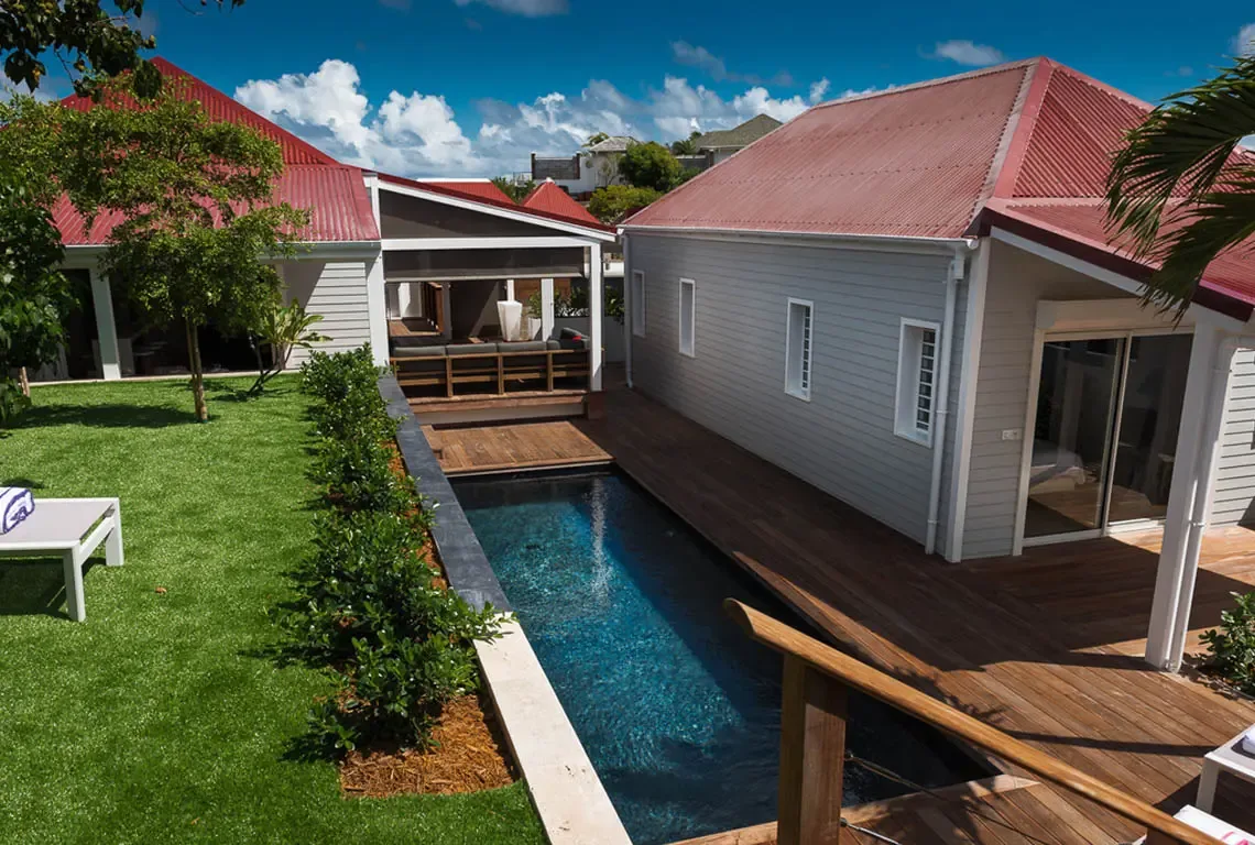 A sleek lap pool in dark blue tile anchors the courtyard, flanked by warm wooden decking and a manicured lawn with white lounger. Red corrugated roofs crown the pavilions while a covered lanai with bar invites shaded socializing—indoor-outdoor luxury.