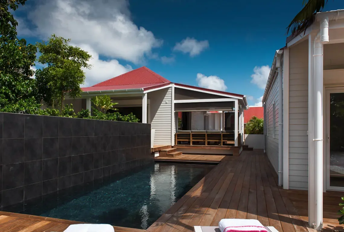 A dark-tiled lap pool reflects the white pavilion with red corrugated roof. Two white loungers await on the wooden deck beside a high gray privacy wall. Blue sky and tropical greenery frame this contemporary Caribbean retreat.