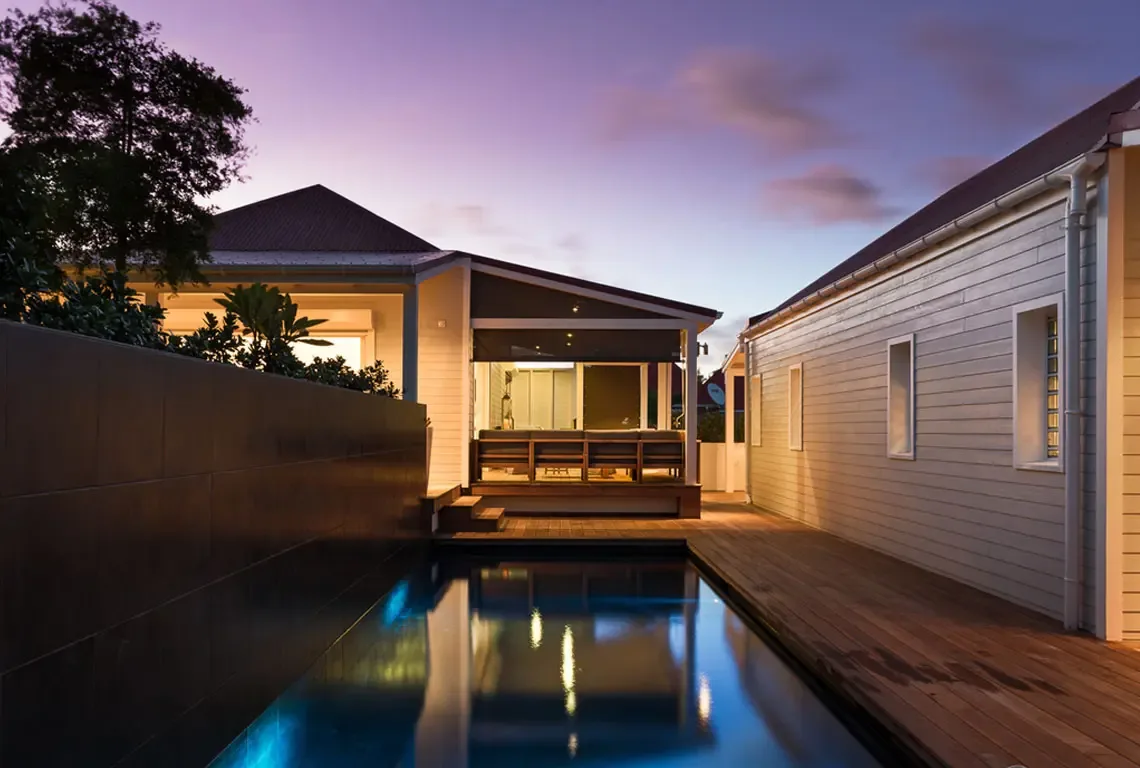 At dusk, the illuminated lap pool glows against warm wooden decking. Modern architecture with gabled roofs and dark tiled walls creates a private sanctuary where indoor comfort meets outdoor elegance—twilight magic by the pool.