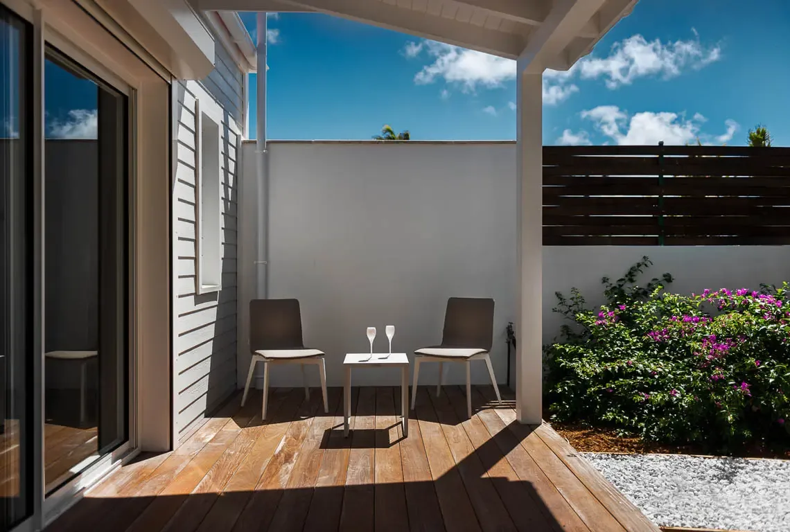 Two modern chairs and a small table with glasses sit on a wood-slat deck beneath a white overhang. Bright pink bougainvillea blooms beside a low white wall while blue sky and scattered clouds complete this intimate terrace for morning coffee or sunset drinks.