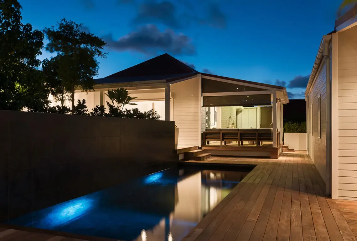 White siding and a dark hip roof define this modern pavilion at dusk. A covered veranda with built-in seating overlooks the illuminated pool, while warm interior lights and glowing blue underwater create a dramatic evening ambiance—perfect for night swims and cocktails.